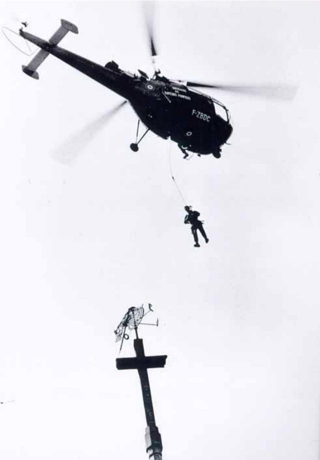 Suspendu au treuil de l'Alouette 3 F-ZBDC de la Brigade des Sapeurs Pompiers de Paris, le sergent Fraïoli, armé de pinces coupantes, s'apprête à sectionner le support du coq (5 mai 1972) - Photo DR 
